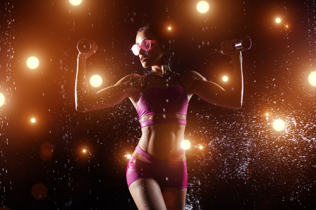 Beautiful caucasian sportswoman posing with dumbbells on a black background in spray of water. Over power concept for poster or bannerの写真素材