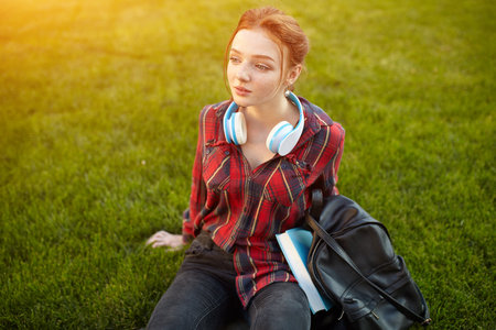 young beautiful student wearing a checkered shirt headphones and a backpack resting on the street between classes. Beautiful redhead girlの写真素材