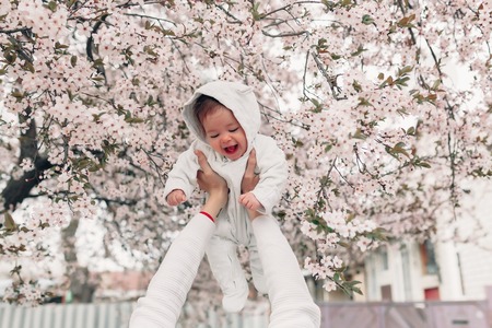 Portrait of happy joyful child in white clothes over tree flowers blossom background. Family playing together outside. Mom cheerfully hold little daughter Newborn spring conceptの写真素材