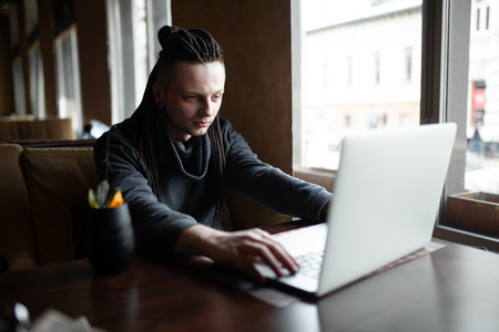 Young Businessman with dreadlock having doing his work in cafe near the big window with laptop. Youth culture. Neformal guy. Remote works. IT specialistの写真素材