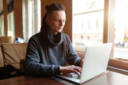 Young Businessman with dreadlock having doing his work in cafe near the big window with laptop. Youth culture. Neformal guy. Remote works. IT specialistの写真素材