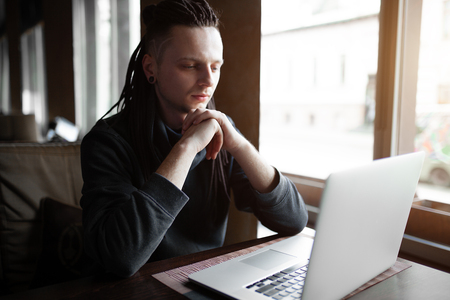 Young Businessman with dreadlock having doing his work in cafe near the big window with laptop. Youth culture. Neformal guy. Remote works. IT specialistの写真素材