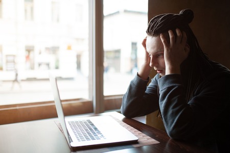 Young confused Businessman with dreadlock having doing his work in cafe near the big window with laptop. Youth culture. Neformal guy. Remote works. IT specialistの写真素材