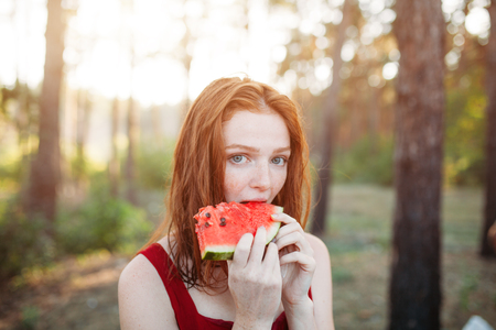 Happy young redhair woman eating watermelon on the nature. Youth lifestyle. Happiness, joy, holiday, beach, summer concept.の写真素材