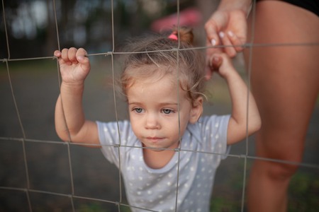 Toned portrait of Sad little girl with her mothe looks through wire fence Summer dayの写真素材