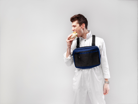 Young stylish male worker with chest rig bag is eating a tasty juicy burger on a white background Isolated on the white backgroundの写真素材