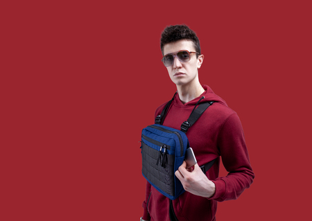 Young handsome guy in sunglasses and red hoodie with a chest rig bag posing in the studio on a red background Model shootingの写真素材