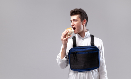 Young stylish male worker with chest rig bag is eating a tasty juicy burger on a white background Isolated on the white backgroundの写真素材