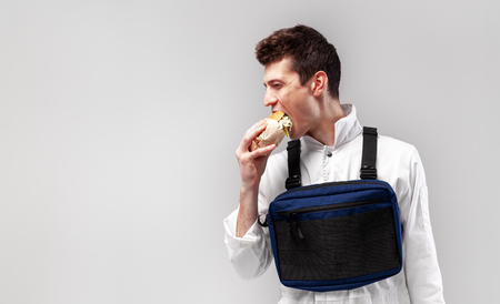 Young stylish male worker with chest rig bag is eating a tasty juicy burger on a white background Isolated on the white backgroundの写真素材