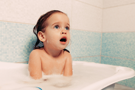 Fun cheerful happy toddler baby taking a bath playing with foam bubbles. Little child in a bathtub. Smiling kid in bathroom on blue background. Hygiene and health careの写真素材