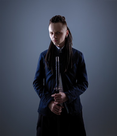 Young man dressed in the white shirt and dark blue jacket with dreadlocks holding a samurai sword. Glamour photo. Studio shooting. business man with Japanes swordの写真素材