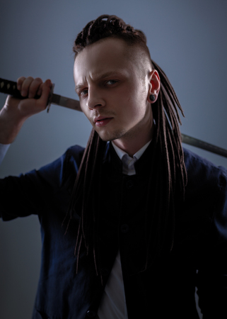Young man dressed in the white shirt and dark blue jacket with dreadlocks holding a samurai sword. Glamour photo. Studio shooting. business man with Japanes swordの写真素材