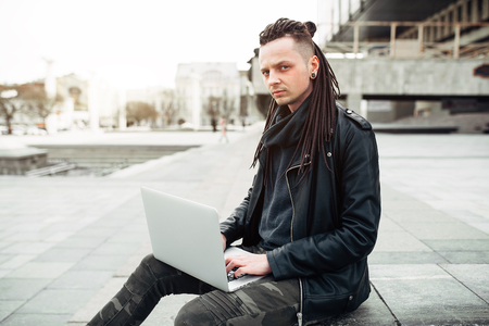 Young fashionable caucasian man with dreadlocks dressed in leather jacket sitting on the stairs using laptop. Remote works IT specialistの写真素材