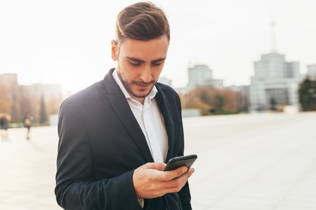 Millennial businessman looks into the screen of his mobile phone. Close-up portrait. Young successful, stylish business man walks on a city street, watching social networks in his smartphone.の写真素材