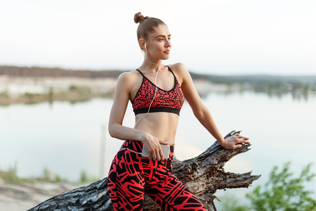 A beautiful brunette girl in stylish sports clothes is resting after a sport active workout near the lake. Listens to music on headphones with a smartphone in handsの写真素材