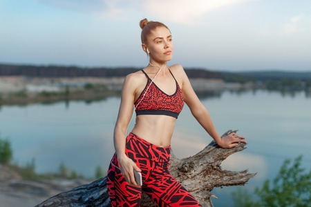 A beautiful brunette girl in stylish sports clothes is resting after a sport active workout near the lake. Listens to music on headphones with a smartphone in handsの写真素材