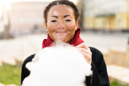 A beautiful young girl of African ethnicity with vitiligo isolated eating cotton candy and smiling. Travel and leisureの写真素材