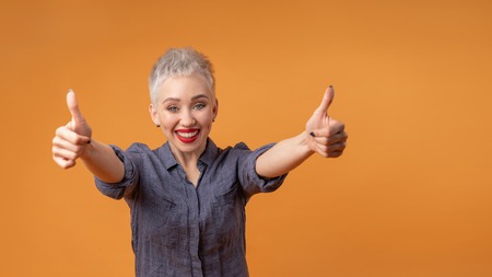 Portrait of young girl with blond short hairstyle looking at camera and laugh showing thumbs up on two hands on orange background with copy space. Woman surprised and smiling. positive emotionの写真素材