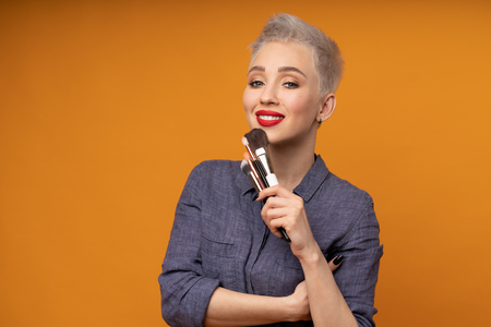 Close up portrait makeup artist. Make up courses. Concept of self visage masterclasses. Woman hold makeup brushes on the hands. Studio shot on the orange color background. Positive people smilingの写真素材