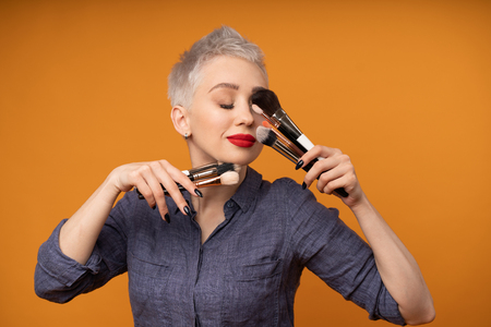 Close up portrait makeup artist. Make up courses. Concept of self visage masterclasses. Fashion professional. Woman hold makeup brushes on the hands. Studio shot on the orange color backgroundの写真素材