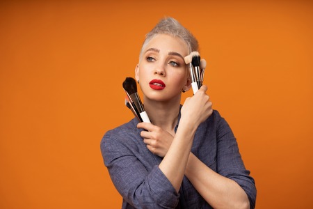 Close up portrait makeup artist. Make up courses. Concept of self visage masterclasses. Fashion professional. Woman hold makeup brushes on the hands. Studio shot on the orange color backgroundの写真素材