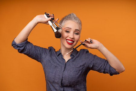 Close up portrait makeup artist. Make up courses. Concept of self visage masterclasses. Fashion professional. Woman hold makeup brushes on the hands. Studio shot on the orange color backgroundの写真素材