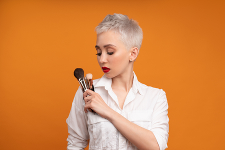 Close up portrait makeup artist. Make up courses. Concept of self visage masterclasses. Fashion professional. Woman hold makeup brushes on the hands. Studio shot on the orange color backgroundの写真素材