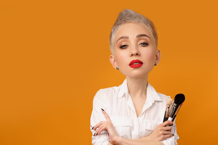 Close up portrait makeup artist. Make up courses. Concept of self visage masterclasses. Fashion professional. Woman hold makeup brushes on the hands. Studio shot on the orange color backgroundの写真素材