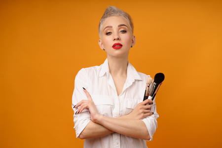 Close up portrait makeup artist. Make up courses. Concept of self visage masterclasses. Fashion professional. Woman hold makeup brushes on the hands. Studio shot on the orange color backgroundの写真素材