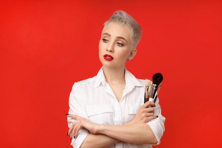 Close up portrait makeup artist. Make up courses. Concept of self visage masterclasses. Fashion professional. Woman hold makeup brushes on the hands. Studio shot on the red color backgroundの写真素材