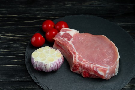 Top view of one pieces raw pork chop steaks with cherry tomatoes and garlic on a black stone cutting...