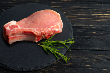 Top view of one pieces raw pork chop steaks with rosemary on a black stone cutting board. Wooden table.の写真素材