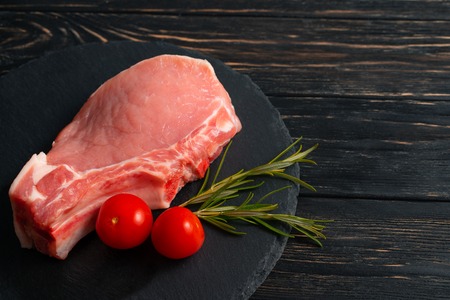 Top view of one pieces raw pork chop steaks with cherry tomatoes and rosemary on a black stone cutting board.. Wooden table.の写真素材