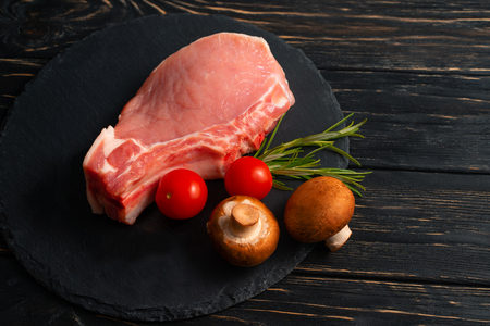 Top view of one pieces raw pork chop steaks with cherry tomatoes Champignon mushrooms and rosemary on a black stone cutting board.. Wooden table.の写真素材