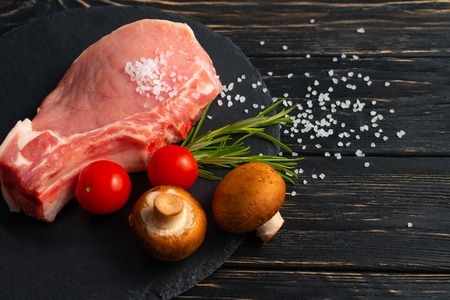 Top view of one pieces raw pork chop steaks with cherry tomatoes Champignon mushrooms sea salt and rosemary on a black stone cutting board.. Wooden table.の写真素材