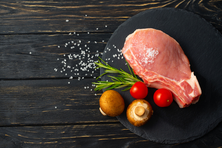 Top view of one pieces raw pork chop steaks with cherry tomatoes Champignon mushrooms sea salt and rosemary on a black stone cutting board.. Wooden table.の写真素材
