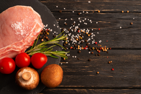 Top view of one pieces raw pork chop steaks with cherry tomatoes Champignon mushrooms sea salt red black pepper and rosemary on a black stone cutting board.. Wooden table.の写真素材