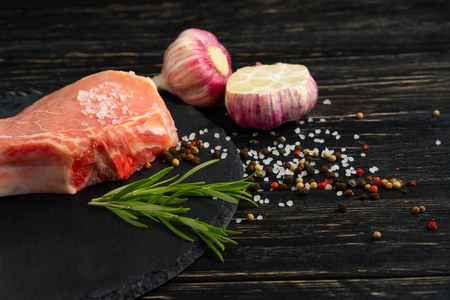 Top view of one pieces raw pork chop steaks with garlic sea salt red black pepper and rosemary on a black stone cutting board.. Wooden table.の写真素材