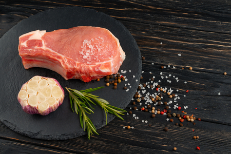 Top view of one pieces raw pork chop steaks with garlic sea salt red black pepper and rosemary on a black stone cutting board.. Wooden table.の写真素材