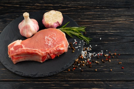 Top view of one pieces raw pork chop steaks with garlic sea salt red black pepper and rosemary on a black stone cutting board.. Wooden table.の写真素材