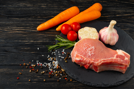 Top view of one pieces raw pork chop steaks with cherry tomatoes carrot garlic sea salt red black pepper and rosemary on a black stone cutting board.. Wooden table.の写真素材