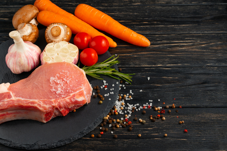 Top view of one pieces raw pork chop steaks with Champignon mushrooms cherry tomatoes carrot garlic sea salt red black pepper and rosemary on a black stone cutting board.. Wooden table.の写真素材