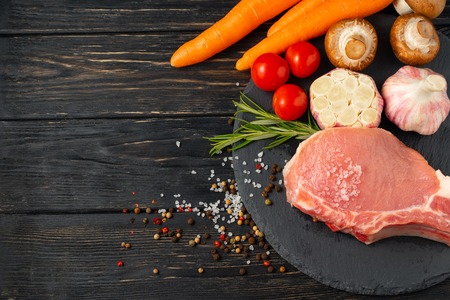 Top view of one pieces raw pork chop steaks with Champignon mushrooms cherry tomatoes carrot garlic sea salt red black pepper and rosemary on a black stone cutting board.. Wooden table.の写真素材