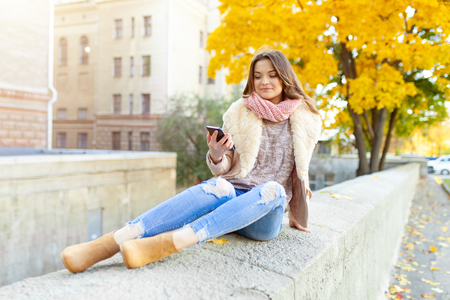 Autumn season A beautiful young brunette girl walks through the city in the rays of warm sunlight. Enjoys warm weather. Holding smartphone in the hand. Texting messageの写真素材