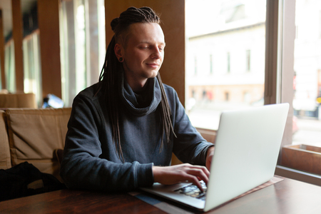 Young Businessman with dreadlock having doing his work in cafe near the big window with laptop. Youth culture. Neformal guy. Remote works. IT specialistの写真素材