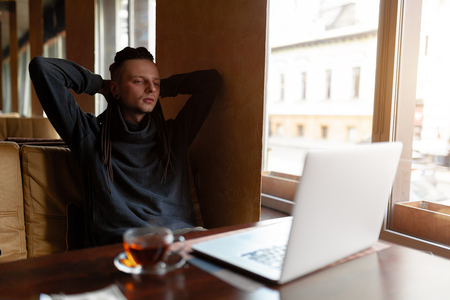 Young Businessman with dreadlock having doing his work in cafe near the big window with laptop. Youth culture. Neformal guy. Remote works. IT specialistの写真素材