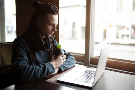 Young Businessman with dreadlock having doing his work in cafe near the big window with laptop. Youth culture. Neformal guy. Remote works. IT specialistの写真素材