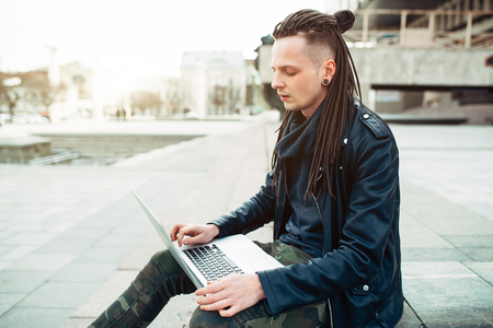Young fashionable caucasian man with dreadlocks dressed in leather jacket sitting on the stairs using laptop. Remote works IT specialistの写真素材