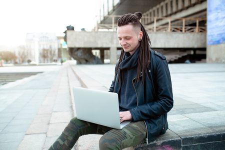 Young fashionable caucasian man with dreadlocks dressed in leather jacket sitting on the stairs using laptop. Remote works IT specialistの写真素材