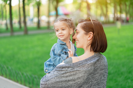 A little Caucasian girl dressed in a denim jacket sits in her mother's arms. Mom and daughter gulf in the park on a hot summer day lifestyleの写真素材